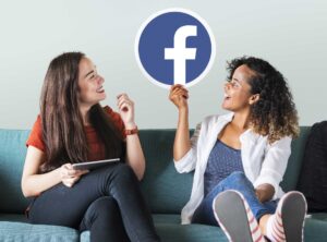 Duas mulheres sentadas em um sofá e segurando um ícone do Facebook.